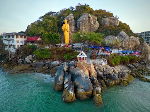 a coastal view in Hua Hin with a towering golden Buddha statue standing on a rocky outcrop overlooking the sea