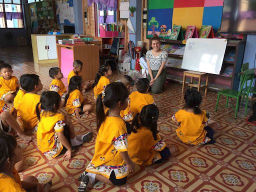 A classroom with a group of children in yellow shirts sitting on the floor watching a teacher who is teaching animal names in English using drawings on a whiteboard
