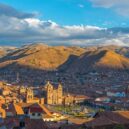 A panoramic view of Cusco, Peru, with terracotta rooftops and the Cathedral of Cusco in the foreground, surrounded by green mountains under a blue sky with scattered clouds.