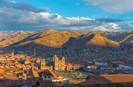 A panoramic view of Cusco, Peru, with terracotta rooftops and the Cathedral of Cusco in the foreground, surrounded by green mountains under a blue sky with scattered clouds.